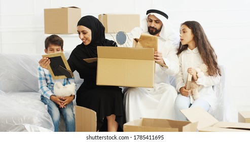 Arabian Married Couple In Traditional Middle Eastern Clothes Siting On Couch And Unpacking Carton Houses. Kids Helping To Parents When Family Moving In New Accomodation. Arabs In Traditional Clothes.