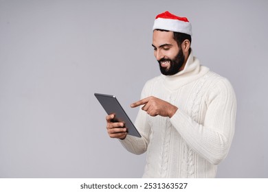 Arabian middle-eastern man in Santa Claus red hat using digital tablet for Christmas shopping online, texting, e-commerce, remote work isolated over grey background - Powered by Shutterstock