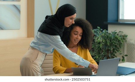 Arabian muslim islamic businesswoman in hijab mentoring new employee African American woman friendly help teach working with laptop in office diverse business women discuss online project write notes - Powered by Shutterstock