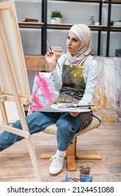 arabian woman with paintbrush and palette looking at canvas on easel