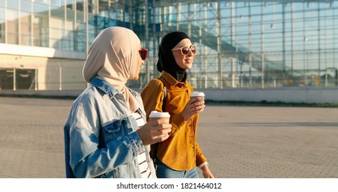 Arabian Young Stylish Women In Traditional Headscarves And Sunglasses Walking Outside At Big Glass Building Of Airport, Talking And Drinking Coffee.