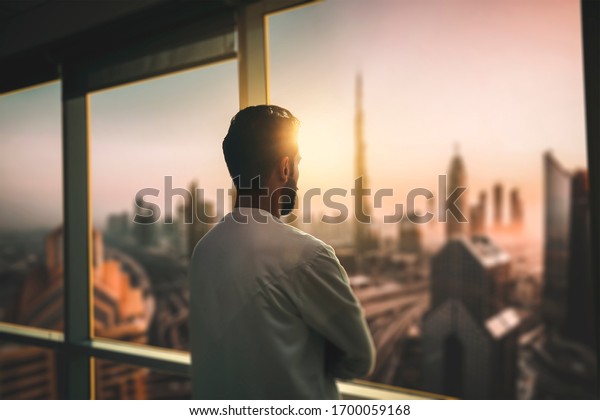 Arabic business man looking out through the\
office balcony seen through glass window. arab young man looking at\
Dubai city through hotel\
window.