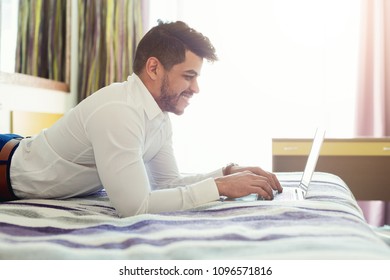 Arabic businessman lying on bed in hotel and using laptop. Smiling man working in hotel room and typing on computer keyboard, side view, copy space - Shutterstock ID 1096571816