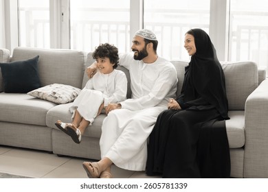 Arabic couple and little son dressed in traditional Muslim attire sit on sofa in living room, smile, look aside, enjoy time together and pleasant weekend rest in modern apartment. Culture, family ties
