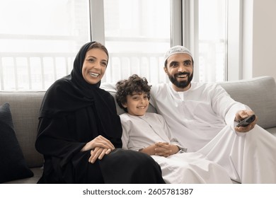 Arabic couple with little son dressed in traditional attire relaxing on couch, holding remote controller switch channels, choosing educational program on internet or movie, spending leisure at home - Powered by Shutterstock - Get 15% off with code: PIKWIZARD15