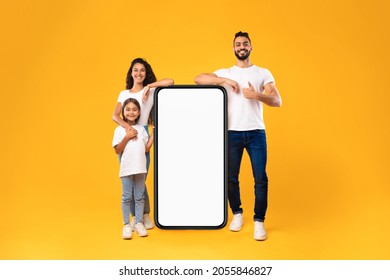 Arabic Family Standing Near Big Smartphone With Blank Screen Advertising Mobile Application Standing Over Yellow Background In Studio. Great App Advertisement. People And Gadget Mockup