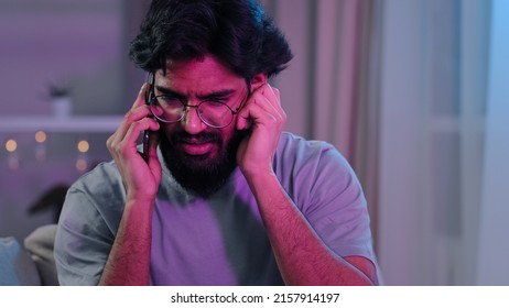 Arabic Indian Bearded Man With Glasses Talking On Phone At House Party Sitting On Sofa Neon Light Hard To Hear Conversation Loud Music Noisy Birthday Celebration Answering Phone Call Trying Listening