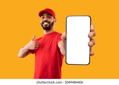 Arabic Male Courier Showing Big Phone With Blank Screen And Pointing Finger Standing On Yellow Background. Studio Shot Of Delivery Man Advertising Mobile Application. Mockup