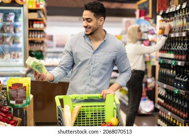 Arabic Male Customer Doing Grocery Shopping Choosing Organic Vegetables Standing With Shop Cart Full Of Food Products In Modern Supermarket. Male Buyer Purchasing Staples In Store