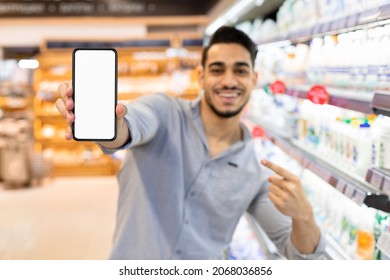 Arabic Male Showing Blank Phone Screen Advertising Grocery Shopping Application Standing In Supermarket. Modern Online Shopping Concept. Selective Focus On Smartphone, Mockup