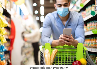 Arabic Man Doing Grocery Shopping Using Smartphone And Wearing Face Mask Standing With Shop Cart In Supermarket. Guy Searching Discounts, Coupons And Sales Using Mobile Shopper Application On Phone