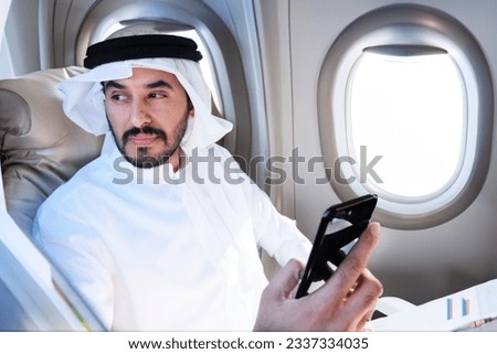 Arabic man flying inside airplane using mobile smartphone looking far away. Middle Eastern in Kandura travelling overseas abroad ideal for holiday vacation or business trip concept