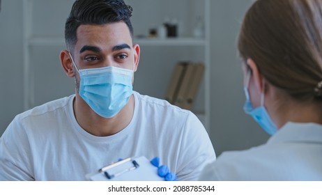 Arabic Man Hispanic Guy Patient Sick Male Wears Medical Protective Mask On Face Talks About Symptoms Of Health Problems To Unrecognizable Doctor Woman Nurse Writing Down Notes, Consultation In Clinic