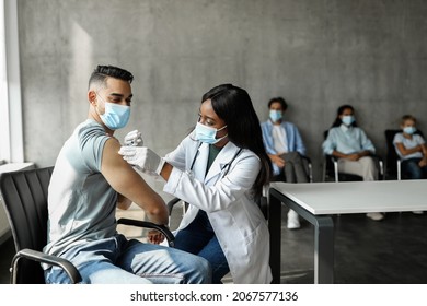 Arabic man patient getting intramuscular shot against COVID-19 at walk-in vaccination site, black woman doctor making injection in shoulder for young man, wearing protective face masks, copy space - Powered by Shutterstock