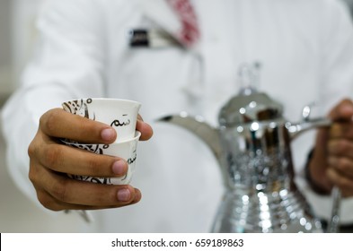 Arabic Man Is Presenting A Cup Of Arabic Coffee (the Translation Of The Text Appears On The Cup: Term Used In Saudi Arabia Means Hospitality Or It Means 