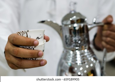 Arabic Man Is Presenting A Cup Of Arabic Coffee (the Translation Of The Text Appears On The Cup: Term Used In Saudi Arabia Means Hospitality Or It Means 