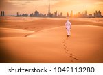 Arabic man with traditional emirates clothes walking in the desert and Dubai skyline in the background