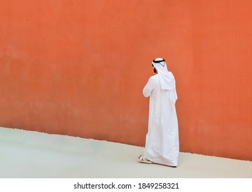 Arabic man with traditional emirates clothes walking outdoors in the street against a orange wall background - Arab ethnicity and muslim religion concept - Copy space for text - Powered by Shutterstock