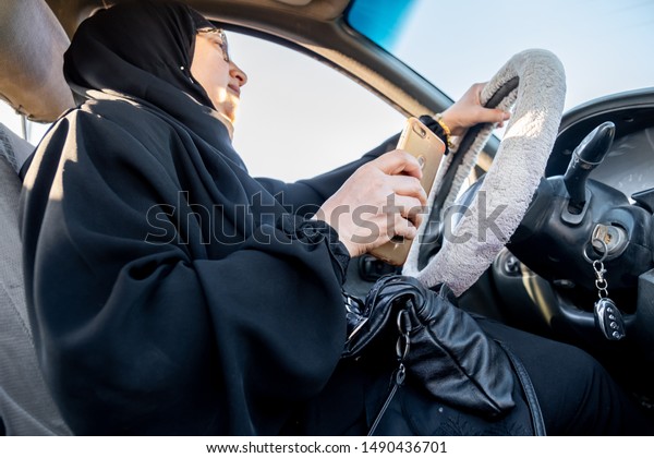 Arabic muslim woman driving a car while using her\
smart phone