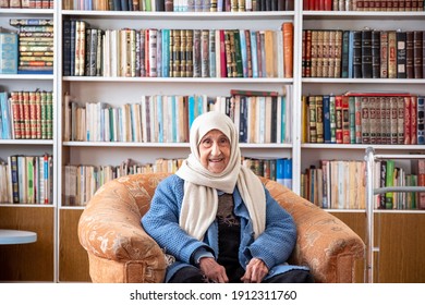 Arabic Old Woman Sitting On Couch In Her Home With A Library Background