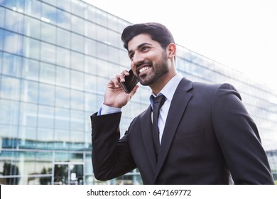 Arabic serious smiling happy successful businessman or worker in black suit with tie and shirt with beard calling with his phone near his ear standing in front of an office building.