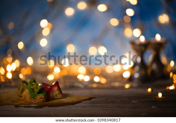 Arabic\
sweets on a wooden surface. Candle holders, night light and night\
blue sky with crescent moon in the\
background.