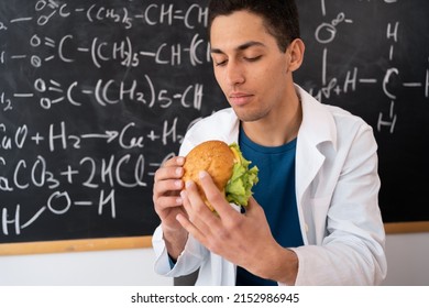 Arabic Young Teacher Eats A Delicious Hamburger In A School Or College Class Sitting At The Table. Hungry Student With Fast Food Lunch Time. Break And Snack On Fatty Hamburger, Unhealthy Food Concept