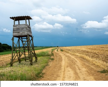 Arable land with hunting observation tower along sandy road with blue sky and clouds.