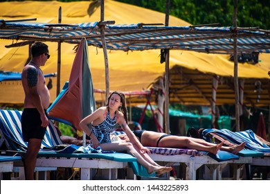 Arambol, Goa, India - 03/04/2019: A Couple At A Beach Shack In The Afternoon. Man Asks His Wife For A Swim In The Sea And She Denies As She Is Not In The Mood. Other Sun Bed, Lady In The Background.