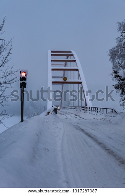 Arc shaped and single lane\
suspension bridge with red light control crossing a river in a\
snowy northern Sweden, a car passing across from the other\
side.