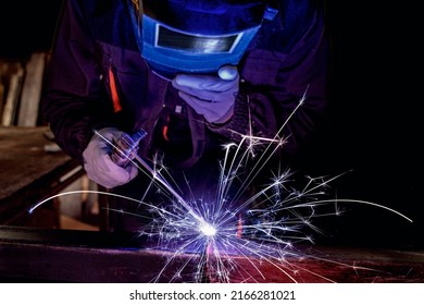 Arc welding. Welder works with electric welding machine. Worker holding electrode clamp, wearing a protective mask, helmet and gloves. Sparks of red-hot electroplasma metal scatter in the dark.