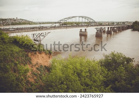 An arch bridge spans over the river, framed by lush trees in the foreground and a city skyline in the background. The natural landscape meets urban architecture in this picturesque scene.