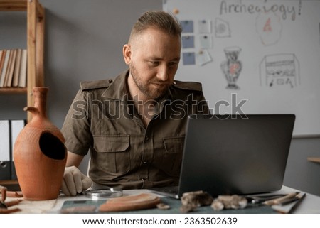 Archaeological office. Male archaeologist doing research, using laptop computer, analyzing of diverse ancient civilization working in his office with old vase. Copy space