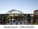 Arched bridge over a river with metal railing in the foreground. Blue sky, trees, and a brick building. Scenic view of bridge and river landscape. Edmund Pettus bridge.