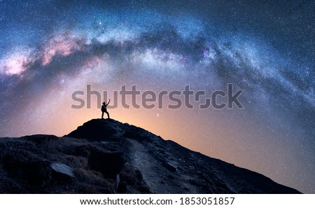 Arched Milky Way and happy man on the mountain at night. Silhouette of guy with raised up arm on the hill, sky with stars, yellow light in Nepal. Galaxy. Space landscape with milky way arch. Travel