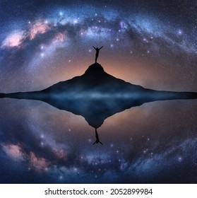 Arched Milky Way And Silhouette Of A Happy Woman On The Mountain Peak At Starry Night. Landscape With Milky Way Arch Reflected In Water, Girl, Sky With Bright Stars, Rock, Lake In Fog. Space. Travel