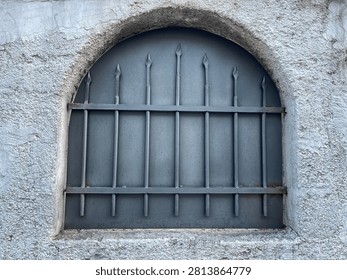 Arched window with iron bars and rough plaster wall texture
Close up of arched window with grey iron security bars set in rough textured plaster wall. Architectural detail background.