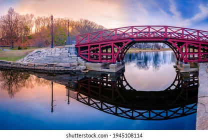 Arching Pink Wooden Bridge and Reflections over the Pond at Elm Park in Worcester, Massachusetts. Tranquil Sunset Landscape over the Mirror Like Lake Water. Asian Style Architecture in American Town.