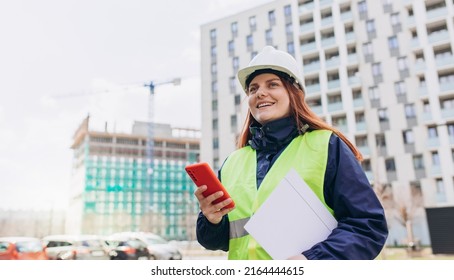 Architect with a blueprints using smartphone at a construction site. Portrait of happy woman constructor wearing white helmet and safety vest outdoors, technology or building concept - Powered by Shutterstock