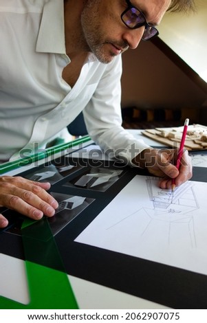 Architect or drafter or engineer or designer, drawing with a pencil in his office on a blueprint or sketch with an architecture model and photos on the table