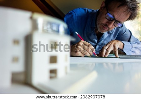 Architect or drafter or engineer or designer, drawing with a pencil in his office on a blueprint or sketch with an architecture model on the blur foreground
