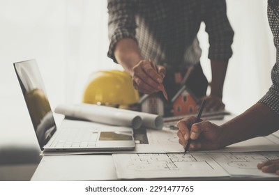 Architect, engineer brainstorm, discuss working plan on village project, house plan, construction on blueprint and view data on laptop computer on office desk.