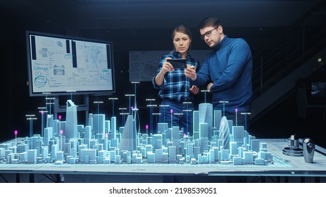 Architect And Engineer Have Discussion, Use Augmented Reality Smartphone To Design Sustainable 3D Megalopolis Model. Futuristic Office With Architectural Designers. Graphical VFX On Image.