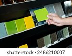 Architect hand choosing and picking green lacquered glass tile on material samples shelf display in the material library. Colorful acrylic sheet collection for interior finishing idea