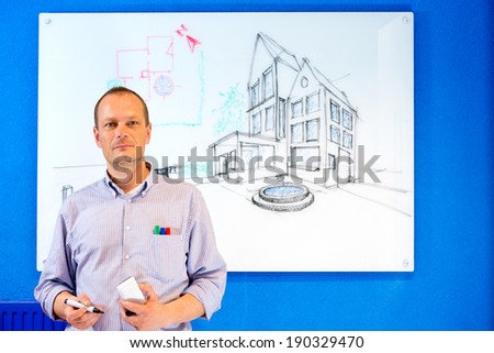 Architect, holding a white board marker, standing in front of a design sketch of a residential structure on the glass board behind him