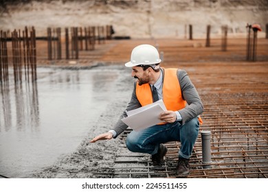 An architect with paperwork in his hands is crouching next to a fresh concrete building foundation and checking on it.