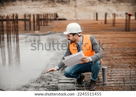 An architect with paperwork in his hands is crouching next to a fresh concrete building foundation and checking on it.