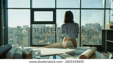 An architect standing by a window overlooking a cityscape, reflecting on project plans with blueprints and tools spread on the desk in a modern workspace