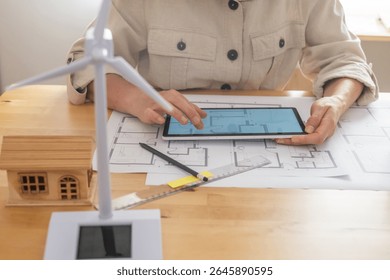 Architect using digital tablet and blueprints, designing sustainable building with wind turbine and solar panel models, focusing on renewable energy and ecological future - Powered by Shutterstock - Get 15% off with code: PIKWIZARD15