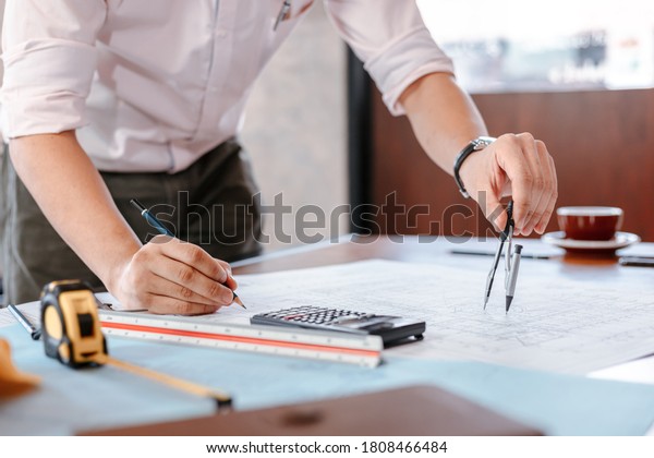 Architect using divider compass working\
on blueprint. blueprints, ruler, calculator, safety hat and book on\
the table. Construction concept. Engineering\
tools